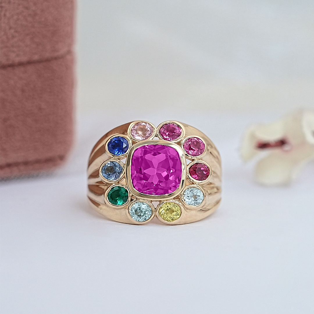 Gold ring with a pink gemstone surrounded by smaller multicolored gems on a light background.